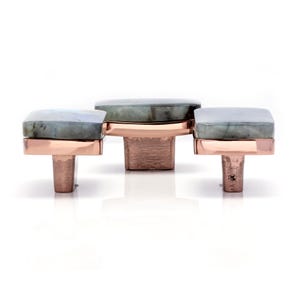 May include: Three cabinet knobs with square, grey-green stone tops and copper-coloured metal bases. The knobs are arranged in a row, with the centre knob slightly larger. The stone has a marbled appearance.