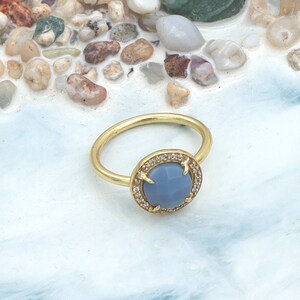 Może przedstawiać: Złoty pierścionek z okrągłym, jasnoniebieskim kamieniem szlachetnym pośrodku. Kamień otoczony jest przez małe, przezroczyste kamienie. Pierścionek leży na białej i niebieskiej powierzchni z małymi kamykami.