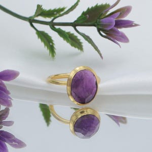 Op de afbeelding: Een gouden ring met een ovale, facetgeslepen paarse edelsteen. De ring heeft een gouden band en zetting. De edelsteen heeft een dieppaarse kleur. De ring is geplaatst tegen een witte achtergrond met paarse bloemen en groene bladeren.