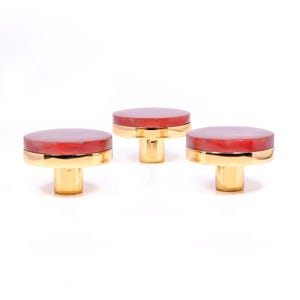 Könnte beinhalten: Drei dekorative Schrankknöpfe mit roter Oberseite und goldfarbenem Sockel. Die Knöpfe haben ein kreisförmiges Design und sind vor einem weißen Hintergrund platziert. Die Knöpfe sind wahrscheinlich aus Metall und für Möbel geeignet.