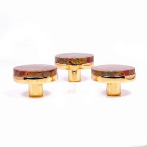 Könnte beinhalten: Drei dekorative Schrankknöpfe mit goldener Oberfläche und farbenfrohen, gemusterten Oberteilen. Die Knöpfe haben eine runde Form und einen kurzen, zylindrischen Sockel. Die Oberteile weisen eine Mischung aus Rot-, Grün- und Gelbtönen auf.