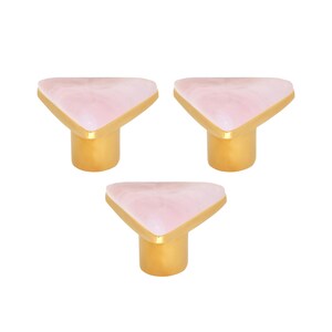 Pode incluir: Três puxadores decorativos para armários. Cada puxador apresenta um topo triangular rosa marmoreado com acabamento dourado, montado numa base cilíndrica dourada. Para móveis e decoração de casa.