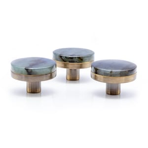 May include: Three cabinet knobs with a round, flat top made of a blue-green stone. The stone is set in a brass-coloured metal band, and the base is a matching brass colour. The knobs are on a white background.