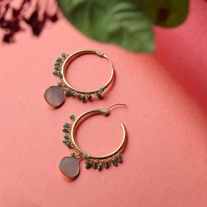 Pode incluir: Um par de brincos de argola dourados. Cada brinco apresenta um pequeno pendente redondo cinza pálido e é adornado com pequenas contas verdes. Os brincos são colocados sobre um fundo rosa.