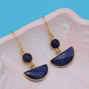 Peut inclure: Une paire de boucles d'oreilles dorées avec un motif de pierres précieuses bleu foncé. Chaque boucle d'oreille comporte une petite pierre ronde au-dessus d'une pierre plus grande en forme de demi-lune. Les boucles d'oreilles sont présentées sur une surface rose sur fond bleu.
