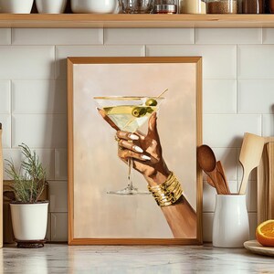 May include: A framed art print of a hand holding a martini glass with olives. The hand is adorned with gold bracelets and rings, and the nails are painted white. The martini glass has a gold rim and is filled with a clear liquid and green olives.