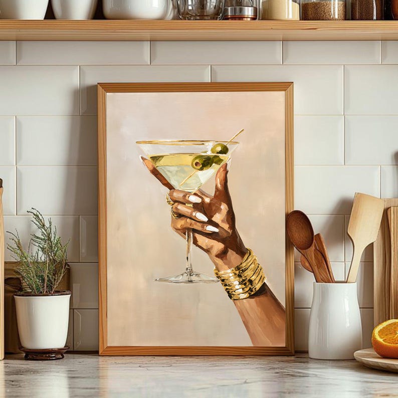 May include: A framed art print of a hand holding a martini glass with olives. The hand is adorned with gold bracelets and rings, and the nails are painted white. The martini glass has a gold rim and is filled with a clear liquid and green olives.