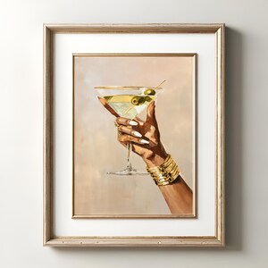 May include: A framed print of a hand holding a martini glass with olives. The hand is adorned with gold bracelets and rings, and the nails are painted white. The martini is garnished with olives on a toothpick.