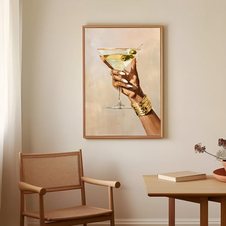 May include: A framed painting depicts a hand holding a martini glass with olives. The hand is adorned with gold bracelets and rings, and the nails are painted white. The martini is a light yellow color, and the background is a soft beige.