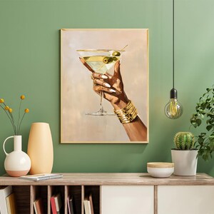 May include: A framed art print depicts a hand holding a martini glass with olives. The hand is adorned with gold bracelets and rings. The background is a soft beige, and the frame is gold. The artwork is displayed on a wooden cabinet.