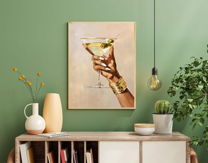 May include: A framed art print depicts a hand holding a martini glass with olives. The hand is adorned with gold bracelets and rings. The background is a soft beige, and the frame is gold. The artwork is displayed on a wooden cabinet.
