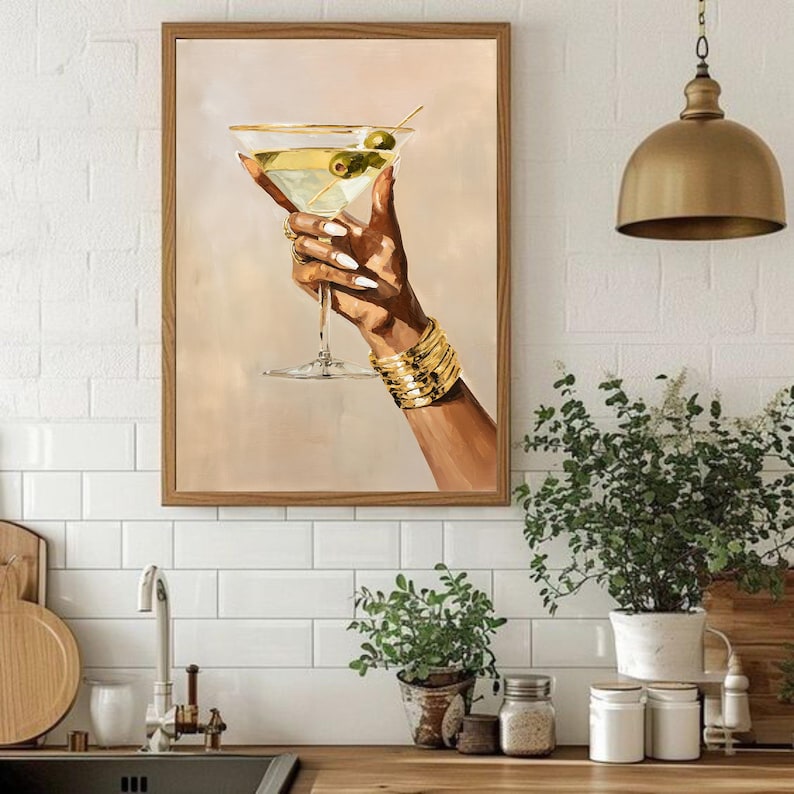 May include: A framed art print depicts a hand holding a martini glass with olives. The hand is adorned with gold bracelets and rings, and the nails are painted white. The martini is filled with a clear liquid and garnished with green olives.