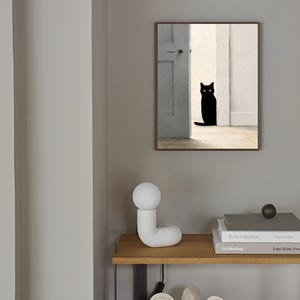 Peut inclure: Une &oelig;uvre encadr&eacute;e repr&eacute;sente un chat noir aux yeux jaunes dans une porte. Le chat est mis en valeur sur un fond de murs blancs et d'une porte grise. L'&oelig;uvre est accroch&eacute;e sur un mur gris au-dessus d'une &eacute;tag&egrave;re en bois avec une lampe blanche et des livres.