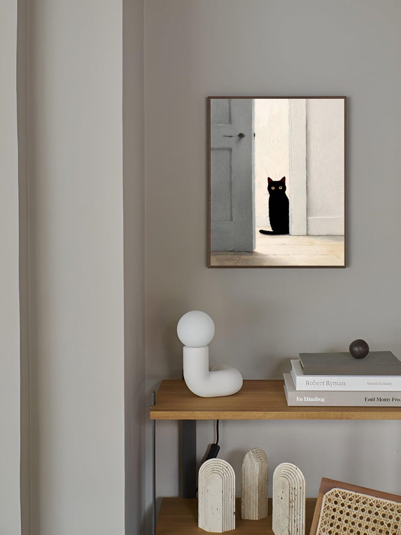 Peut inclure: Une &oelig;uvre encadr&eacute;e repr&eacute;sente un chat noir aux yeux jaunes dans une porte. Le chat est mis en valeur sur un fond de murs blancs et d'une porte grise. L'&oelig;uvre est accroch&eacute;e sur un mur gris au-dessus d'une &eacute;tag&egrave;re en bois avec une lampe blanche et des livres.