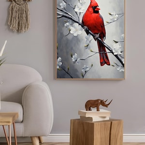 May include: A framed painting of a vibrant red cardinal perched on a branch with white blossoms. The artwork is set against a soft gray background, creating a serene and natural aesthetic. The painting is displayed in a living room setting.