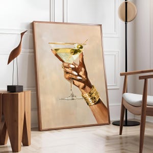 May include: A framed artwork depicts a hand holding a martini glass with olives. The hand is adorned with gold bracelets and rings, and the nails are painted white. The background is a soft beige, and the frame is a warm brown.