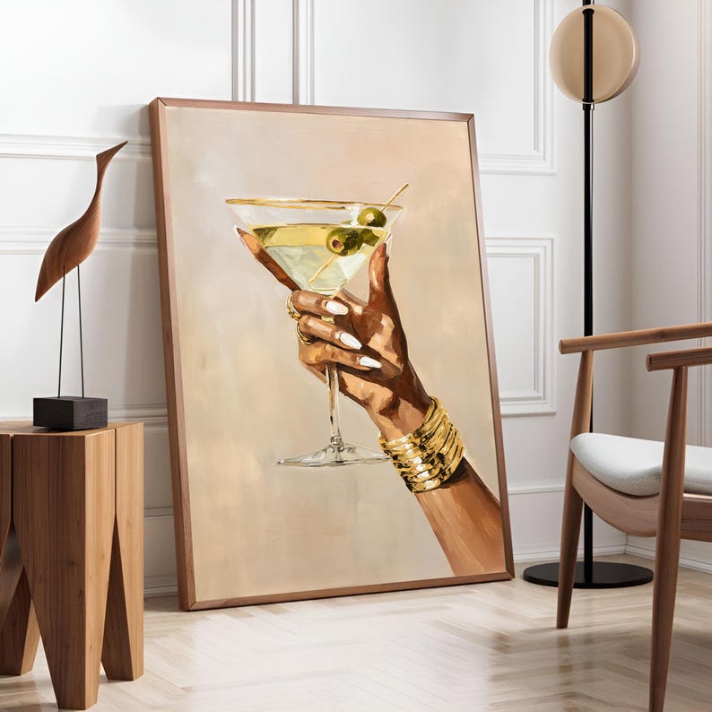 May include: A framed artwork depicts a hand holding a martini glass with olives. The hand is adorned with gold bracelets and rings, and the nails are painted white. The background is a soft beige, and the frame is a warm brown.