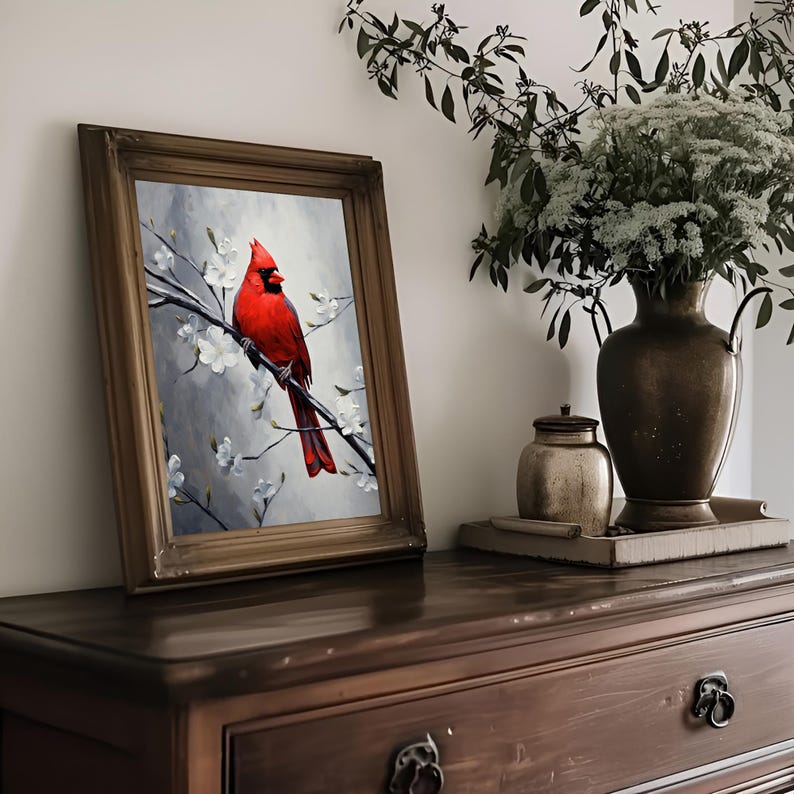 May include: A framed artwork featuring a vibrant red cardinal perched on a branch with white blossoms against a gray backdrop. The artwork is displayed on a dark wooden dresser, with a vase of greenery and a decorative jar in the background.