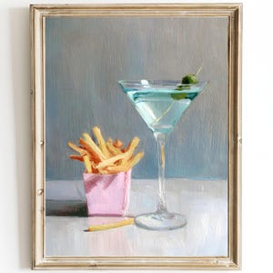 Puede incluir: Pintura enmarcada que representa un cóctel con una aceituna y un recipiente rosa de patatas fritas. La copa de cóctel está llena de un líquido azul. Las patatas fritas son de color marrón dorado. La pintura tiene un fondo azul claro y blanco.