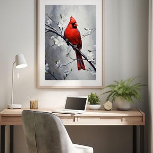 May include: A framed art print of a vibrant red cardinal perched on a flowering branch. The artwork features a gray and white background with white blossoms. The print is displayed above a wooden desk with a laptop, plants, and a desk lamp.