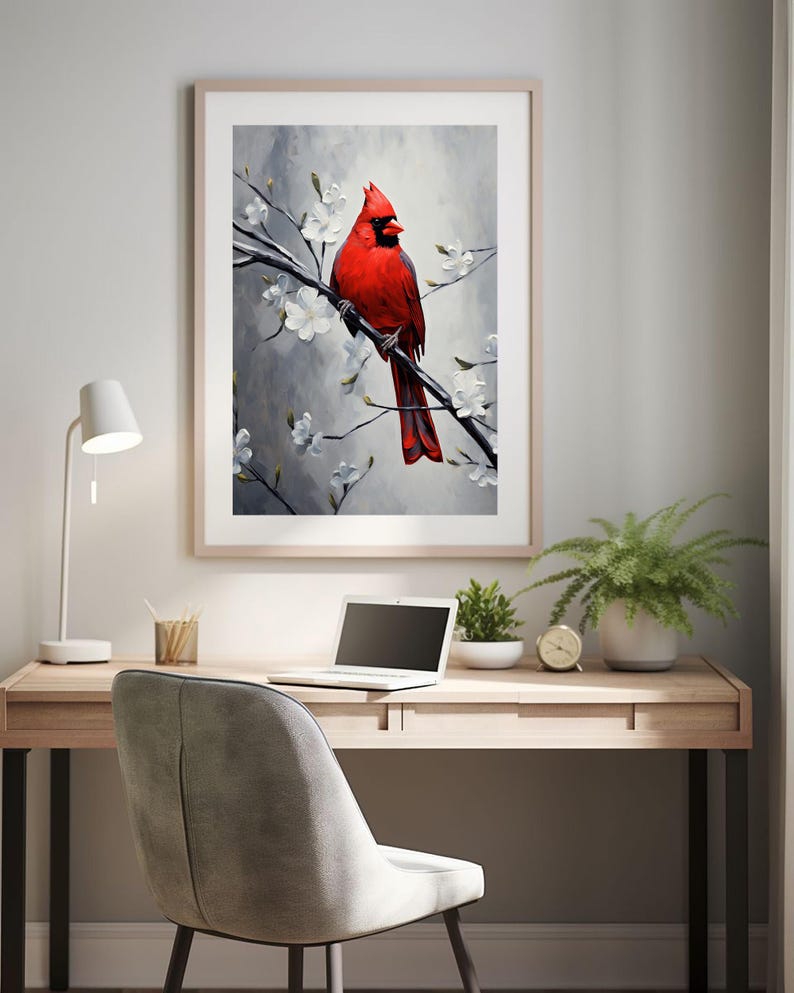 May include: A framed art print of a vibrant red cardinal perched on a flowering branch. The artwork features a gray and white background with white blossoms. The print is displayed above a wooden desk with a laptop, plants, and a desk lamp.