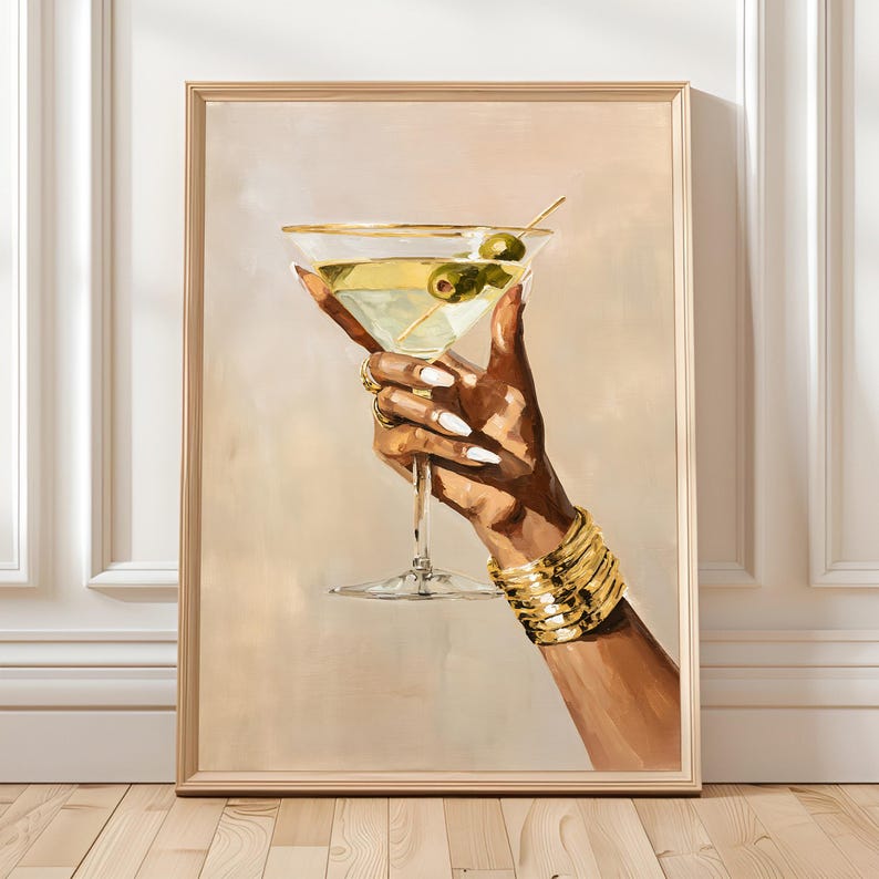 May include: A framed painting depicts a hand holding a martini glass with olives. The hand is adorned with gold jewelry and a gold bracelet. The martini glass has a gold rim and is filled with a yellow liquid. The background is a soft beige.