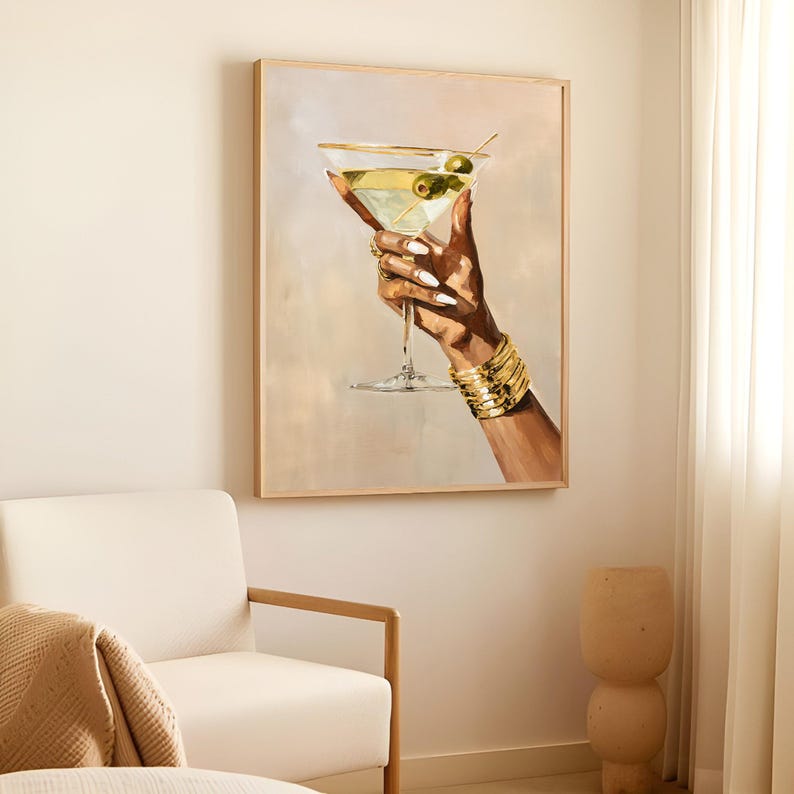 May include: A framed artwork depicts a hand holding a martini glass with olives. The hand is adorned with gold bracelets and rings. The martini glass is filled with a clear liquid and green olives. The background is a soft, neutral color.