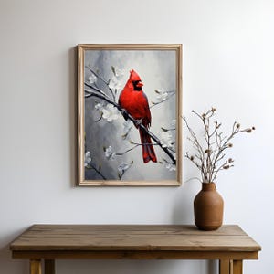 May include: A framed painting of a vibrant red cardinal perched on a branch with white blossoms. The artwork has a soft, painterly style with a gray and white background. The frame is a light wood color. A brown vase with branches sits on a wooden table.