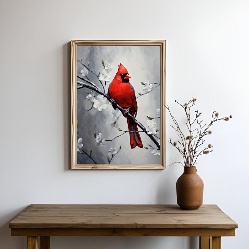 May include: A framed painting of a vibrant red cardinal perched on a branch with white blossoms. The artwork has a soft, painterly style with a gray and white background. The frame is a light wood color. A brown vase with branches sits on a wooden table.