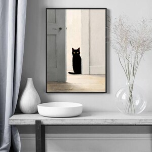 Peut inclure: Impression d'art encadr&eacute;e repr&eacute;sentant un chat noir assis dans une porte. Le chat a les yeux jaunes et se d&eacute;tache sur un fond aux tons neutres. L'&oelig;uvre est expos&eacute;e au-dessus d'un bol et d'un vase blancs sur une table grise.