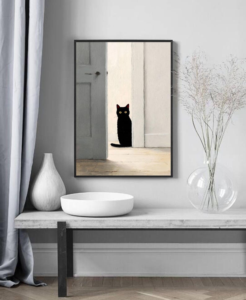 Peut inclure: Impression d'art encadr&eacute;e repr&eacute;sentant un chat noir assis dans une porte. Le chat a les yeux jaunes et se d&eacute;tache sur un fond aux tons neutres. L'&oelig;uvre est expos&eacute;e au-dessus d'un bol et d'un vase blancs sur une table grise.