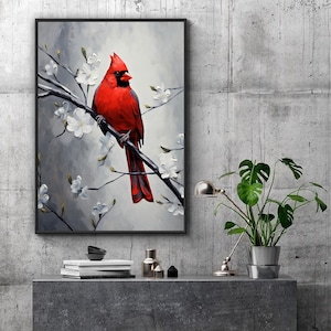 May include: Framed art print featuring a vibrant red cardinal perched on a branch with white blossoms. The bird is set against a soft gray and white background. The artwork is displayed on a gray surface with decorative items and a potted plant.