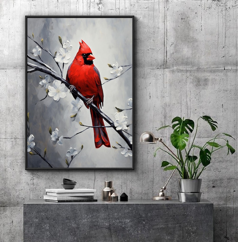 May include: Framed art print featuring a vibrant red cardinal perched on a branch with white blossoms. The bird is set against a soft gray and white background. The artwork is displayed on a gray surface with decorative items and a potted plant.