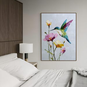 以下が含まれることがあります： ピンクと黄色の花の近くにいるハチドリの油絵。鳥は緑、青、紫の羽を持っています。アートワークは茶色の木製フレームに額装され、明るい灰色の壁に掛けられています。