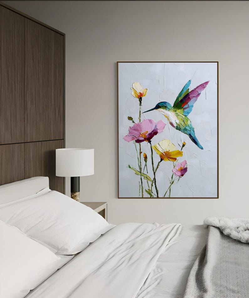 以下が含まれることがあります： ピンクと黄色の花の近くにいるハチドリの油絵。鳥は緑、青、紫の羽を持っています。アートワークは茶色の木製フレームに額装され、明るい灰色の壁に掛けられています。