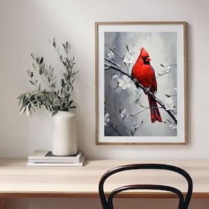 May include: A framed art print featuring a vibrant red cardinal perched on a branch with white blossoms. The artwork has a gray and white background, and the frame is a light wood color. A white vase with greenery sits on a wooden desk.