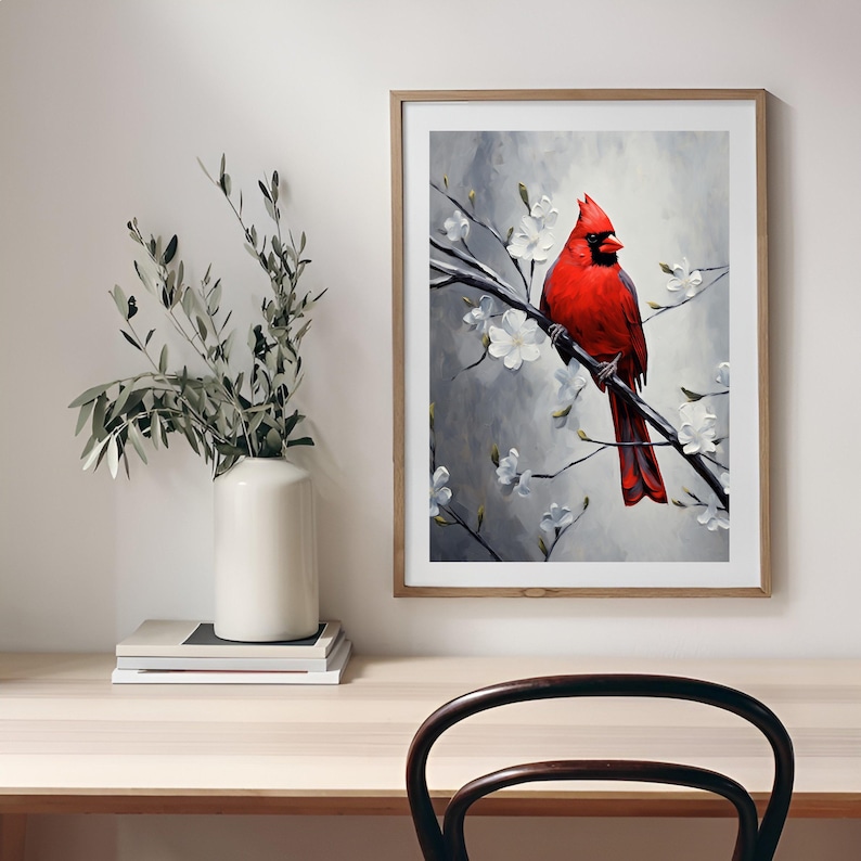 May include: A framed art print featuring a vibrant red cardinal perched on a branch with white blossoms. The artwork has a gray and white background, and the frame is a light wood color. A white vase with greenery sits on a wooden desk.