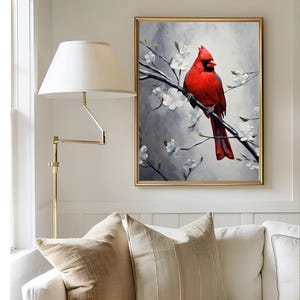 May include: A framed painting of a vibrant red cardinal perched on a branch with white blossoms. The artwork has a gold frame and hangs on a white wall. The painting is a focal point above a white sofa with beige pillows.