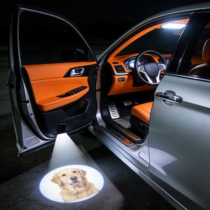 May include: A car door is open, showing an orange interior. A projector shines a light onto the ground, displaying a smiling Golden Retriever. The car's exterior is silver. The interior has black accents and a black steering wheel.