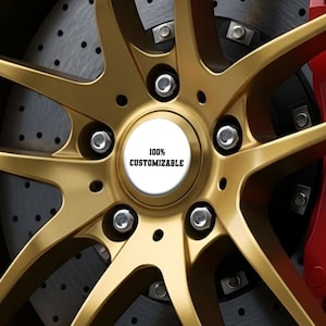 Puede incluir: Primer plano de una llanta de coche dorada con un diseño de múltiples radios. La tapa central es blanca con el texto "100% CUSTOMIZABLE" en negro. La rueda está montada con tuercas plateadas y se ve una pinza de freno roja.