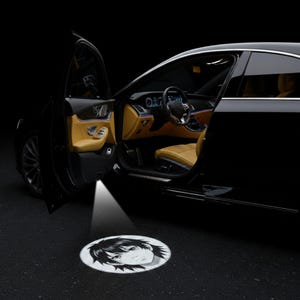 Op de afbeelding: Zwarte auto met de deur open, met een beige interieur. Een cirkelvormig licht projecteert het gezicht van een anime-personage op de grond. Het interieur van de auto is verlicht en de scène speelt zich af tegen een donkere achtergrond.