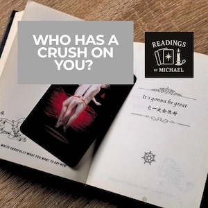 Pode incluir: Um livro aberto com uma carta de tarô com um design vermelho e preto. O texto "WHO HAS A CRUSH ON YOU?" é exibido em um fundo cinza. O livro também tem o texto "It's gonna be great" e "READINGS BY MICHAEL".
