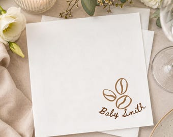 May include: A square white paper napkin featuring a brown printed graphic of three coffee beans and the text Baby Smith in a cursive script font. The napkin is positioned flat, displaying the design clearly in the lower right corner of the item.