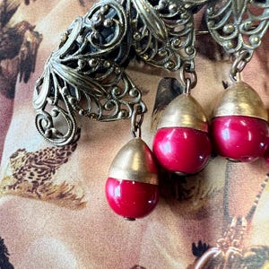 May include: Vintage brass-toned filigree brooch with three dangling red and gold-toned teardrop-shaped pendants. The brooch features intricate openwork designs and is set against a patterned background. The pendants are a vibrant red color.