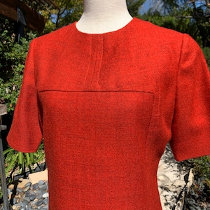May include: A vintage, short-sleeved dress in a vibrant orange-red hue. The dress features a round neckline and a horizontal seam across the chest. The fabric has a subtle textured pattern, and the dress is displayed on a mannequin.