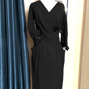 May include: A black dress on a mannequin with a V-neckline and long sleeves. The dress features two small bows and pockets. The mannequin is on a wooden stand. A blue curtain is to the left of the dress.