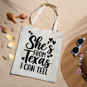 May include: A cream-colored tote bag with the text "She's From Texas I Can Tell" in black script. The bag has two handles and is lying on sand, with seashells and a straw hat with sunglasses nearby.
