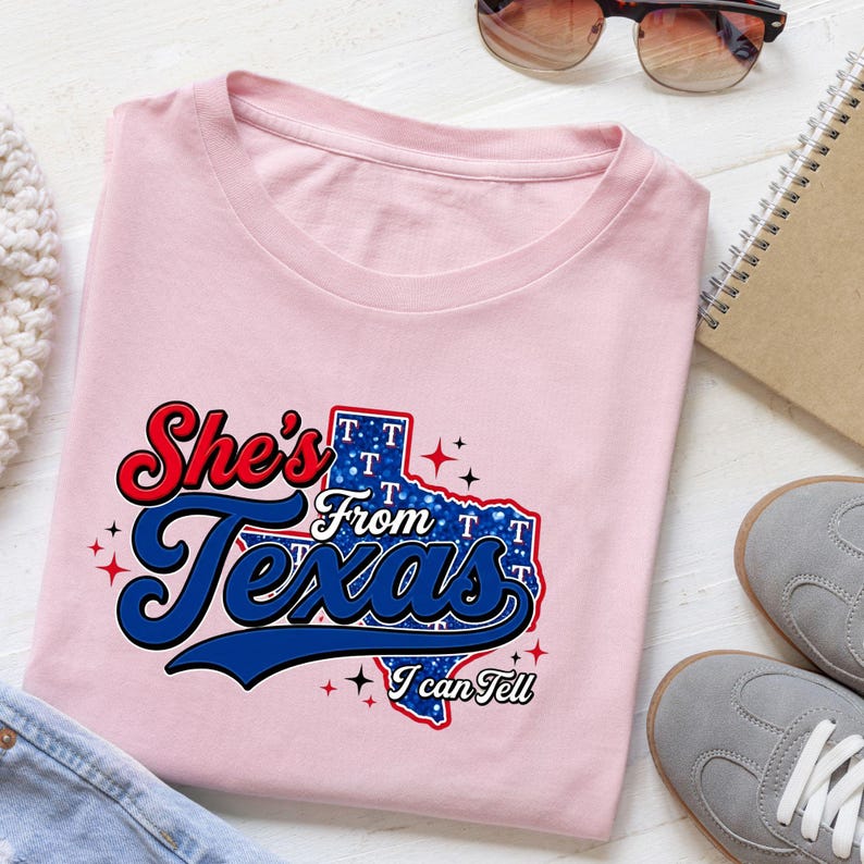 Pode incluir: Uma t-shirt rosa claro com um gr&aacute;fico do estado do Texas preenchido com um padr&atilde;o de glitter azul e a letra T. O texto diz She's From Texas I can Tell numa mistura de fontes vermelhas e azuis com pequenos detalhes de estrelas.