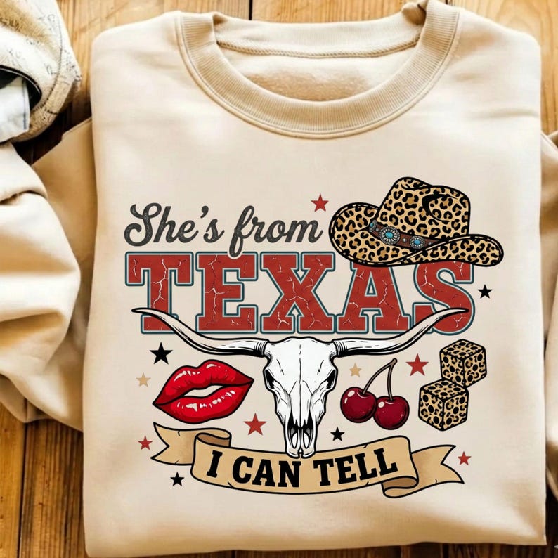 Pode incluir: Uma camisola bege de gola redonda com uma impress&atilde;o gr&aacute;fica com o texto She's from TEXAS I CAN TELL. O design inclui um chap&eacute;u de cowboy com padr&atilde;o de leopardo, um cr&acirc;nio de longhorn branco, l&aacute;bios vermelhos, duas cerejas e dois dados.