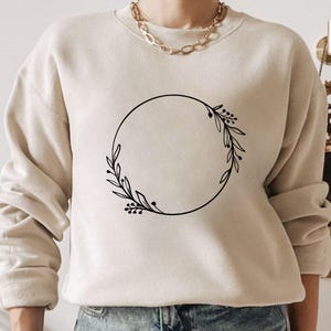 May include: Beige sweatshirt with a black circular floral wreath design. The wreath features leaves and berries. The sweatshirt has long sleeves and a crew neck. A gold chain necklace is visible at the neckline.
